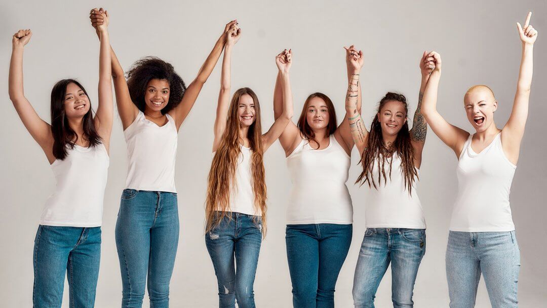 6 Frauen in weißem Shirt und Jeans, die sich an den Händen halten und sie nach oben werfen und lachen.