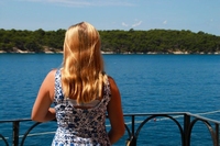 Skin picking Erfahrung Linda klein eine blone Frau, die zum Meer blickt und auf an einer Brüstung steht. Es ist Sommer, sie trägt ein blau-weißes Blumenkleid.